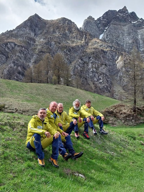 Fünf Bergführer des Ossolalpine-Teams in gelben Jacken sitzen auf einem grünen Almenhang – Sinnbild für Erfahrung und Teamgeist in den Bergen.