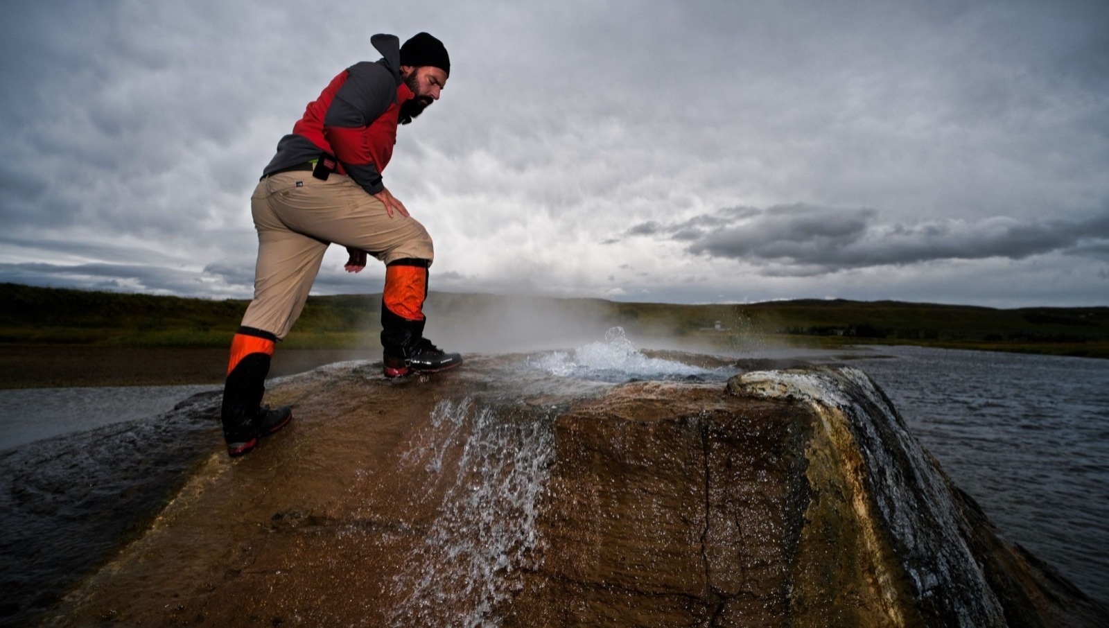 A CoEvolve researcher stands on the edge of a steaming hot spring, observing the water emerging from volcanic rock in a remote, geologically active region.
