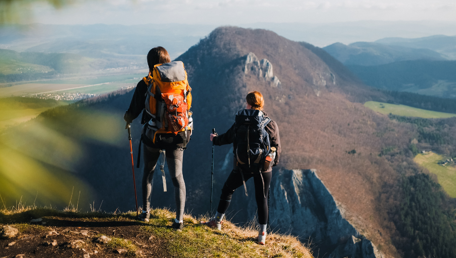 Dos senderistas con mochilas grandes y bastones de trekking contemplan el paisaje desde un mirador montañoso, con colinas boscosas y un valle al fondo.