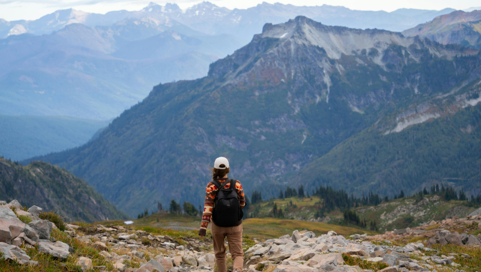 Persona con mochila y gorra caminando por un sendero rocoso, rodeada de montañas cubiertas de bosque y picos escarpados al fondo.