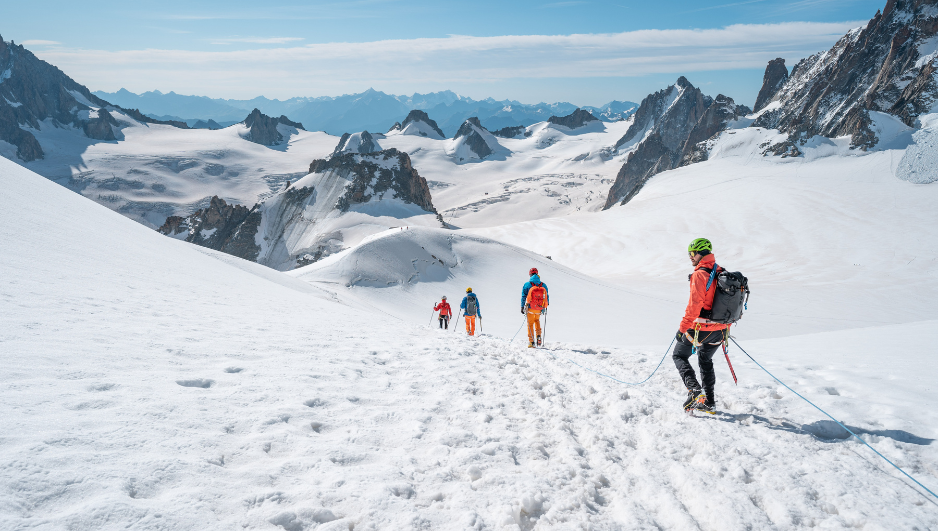 Grupo de alpinistas encordados descendiendo por una ladera nevada en alta montaña, rodeados de picos escarpados y glaciares bajo un cielo azul despejado.