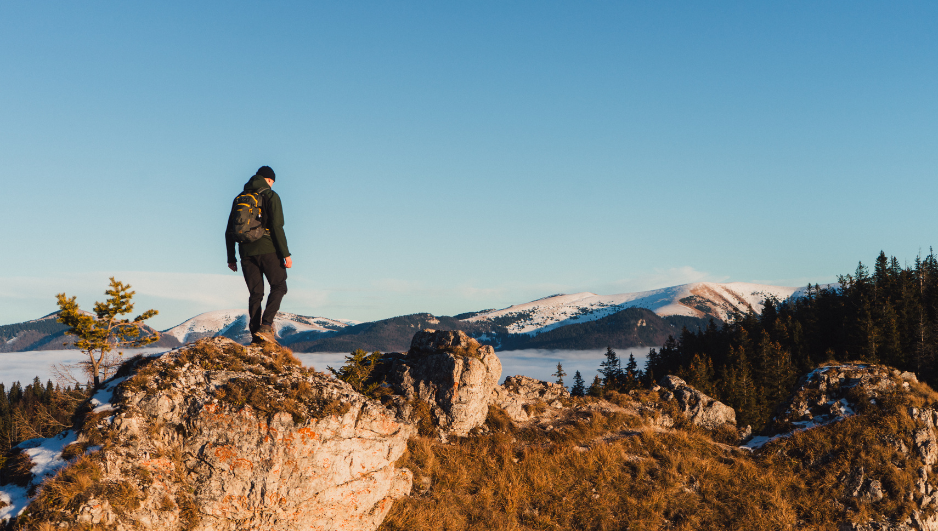 Persona con mochila de senderismo de pie sobre una roca, contemplando un paisaje montañoso nevado bajo un cielo despejado, rodeado de bosques y colinas.