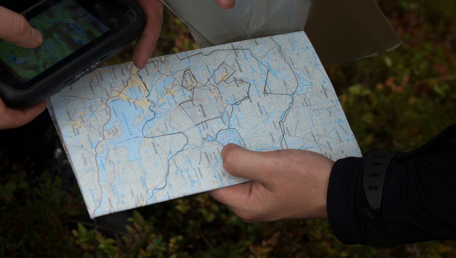 Manos sosteniendo un mapa topográfico detallado al aire libre, con líneas trazadas a mano, junto a un dispositivo GPS con pantalla encendida.