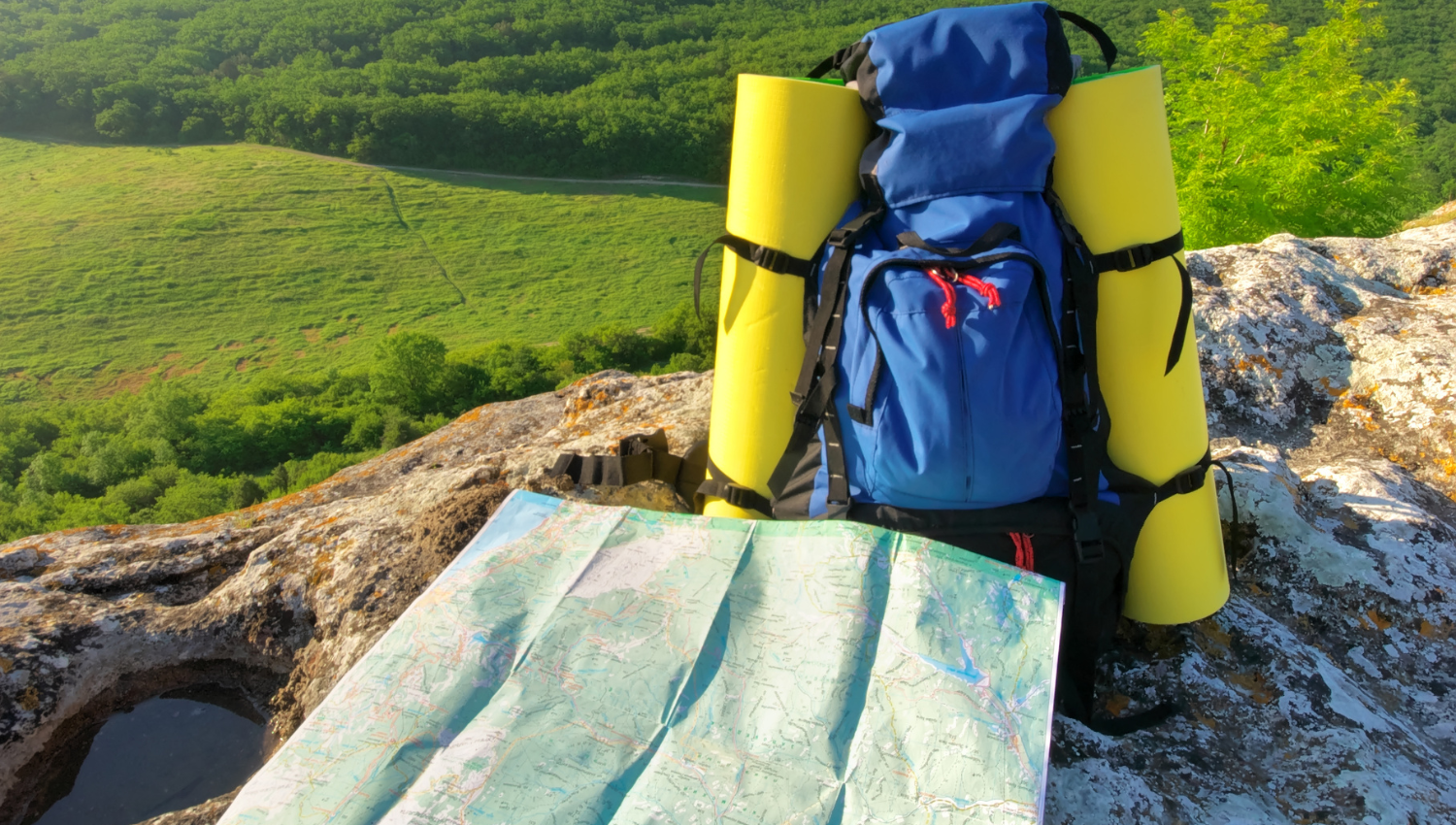 Mochila azul de senderismo con colchonetas amarillas, apoyada sobre una roca, junto a un mapa desplegado con vista panorámica de un valle verde al fondo.