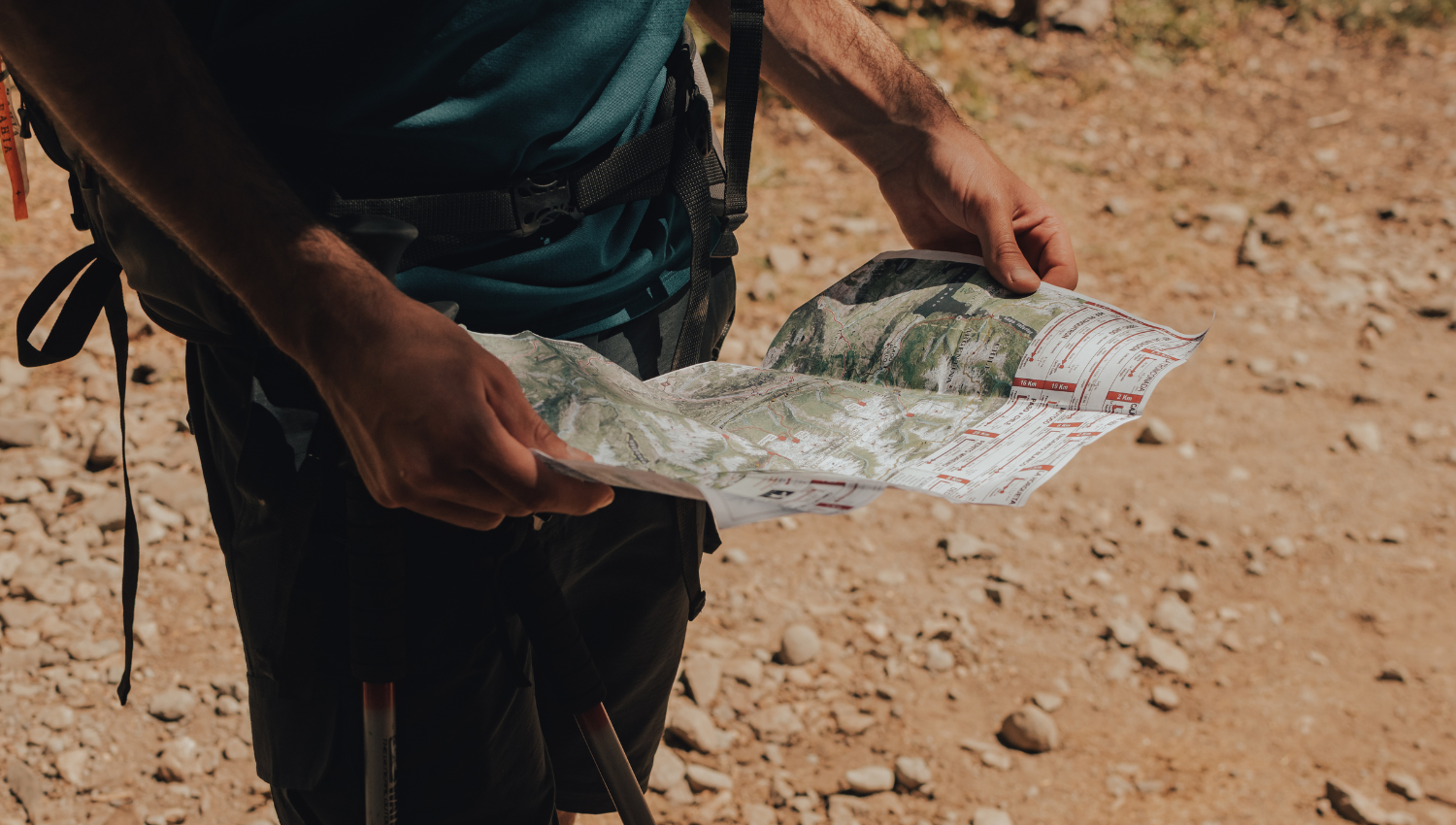 Persona con ropa de senderismo consultando un mapa topográfico al aire libre, en un sendero de tierra rodeado de piedras.