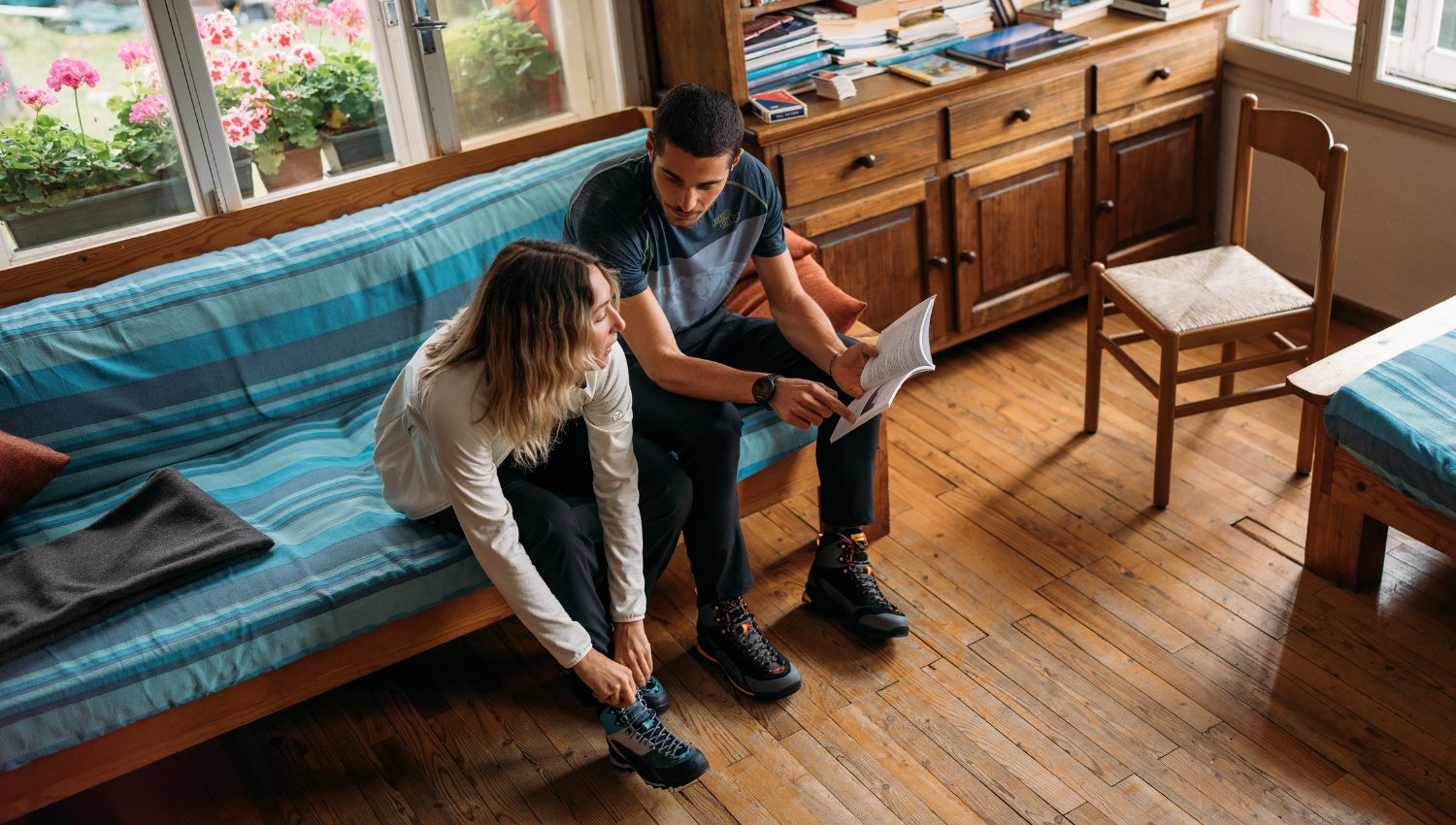 Dos personas sentadas en un sofá con funda azul en una cabaña de montaña; una se está atando las botas de senderismo mientras la otra sostiene una guía abierta y señala algo en la página.