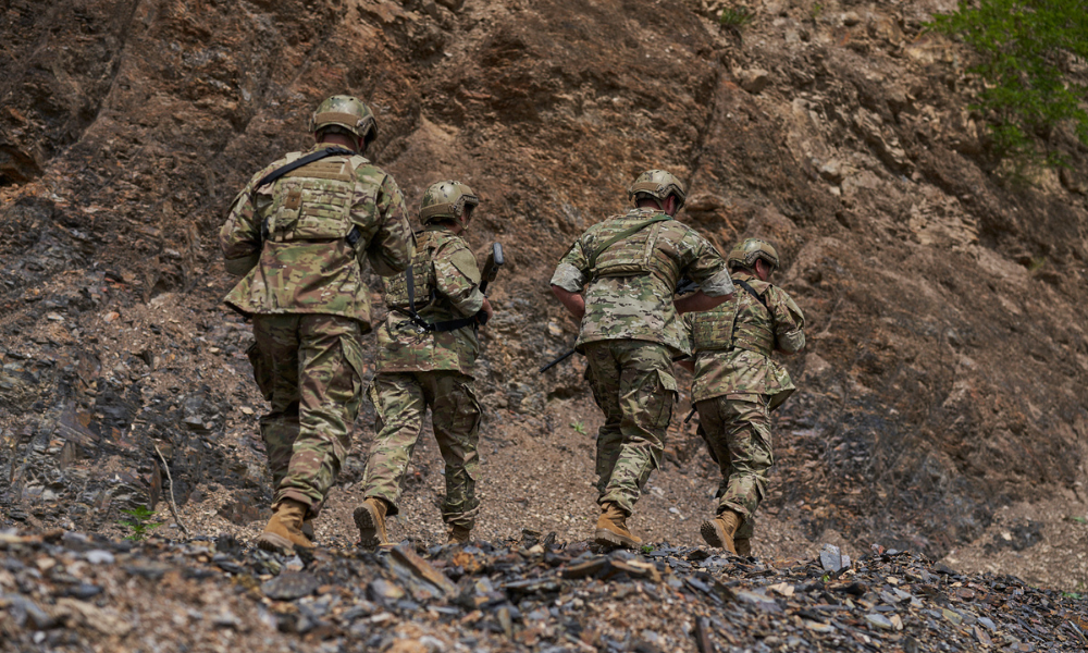 Four soldiers in full camouflage uniform and tactical gear hiking uphill through a rocky terrain, with a steep cliff face beside them.