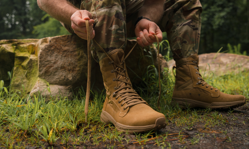 Person in camouflage uniform sitting on a rock and tying the laces of Garmont Tactical tan tactical boots in a forested setting