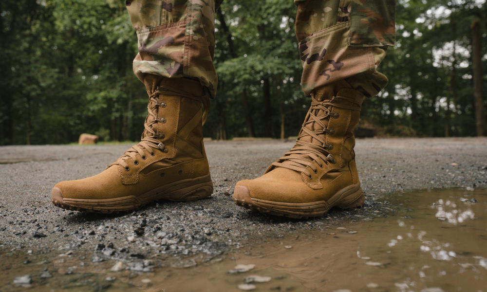 Close-up of Garmont Tactical tan military boots worn with camouflage pants, standing on wet gravel near a puddle in a wooded area