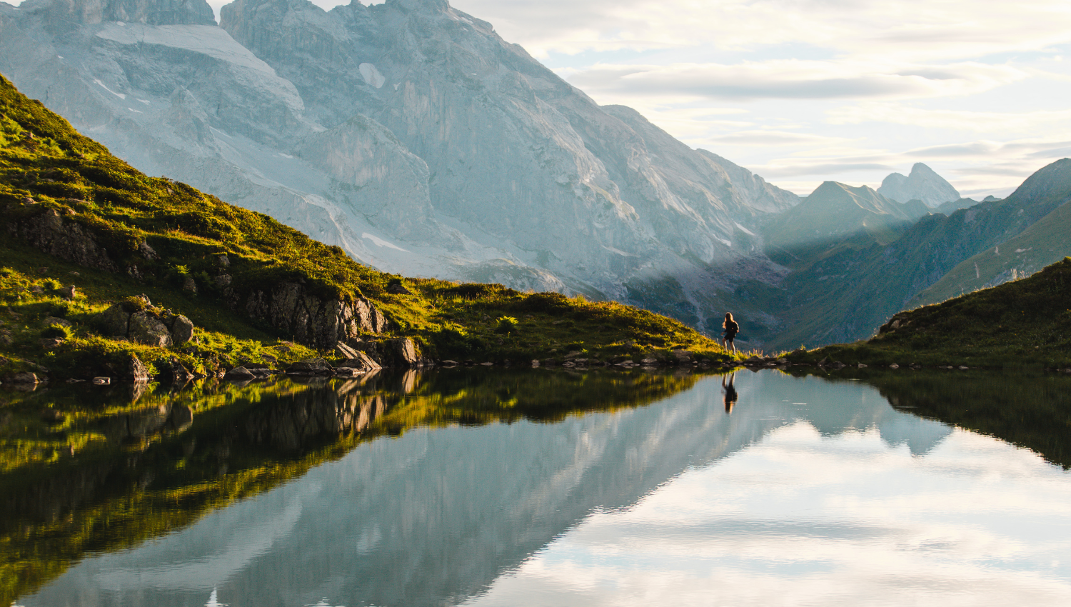 Persona caminando junto a un lago alpino rodeado de montañas, con el paisaje reflejado en el agua
