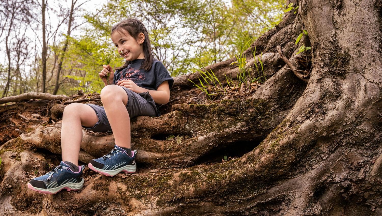 Niña sentada sobre raíces de un árbol grande en el bosque, sonriendo mientras sostiene flores silvestres