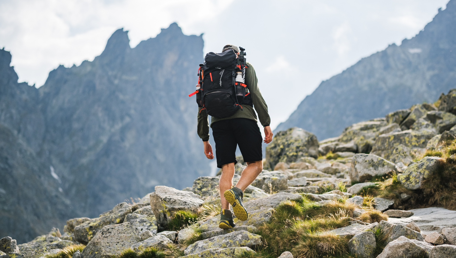 Escursionista che cammina su un sentiero roccioso in alta montagna, con zaino tecnico e scarponi, circondato da cime appuntite e cielo nuvoloso