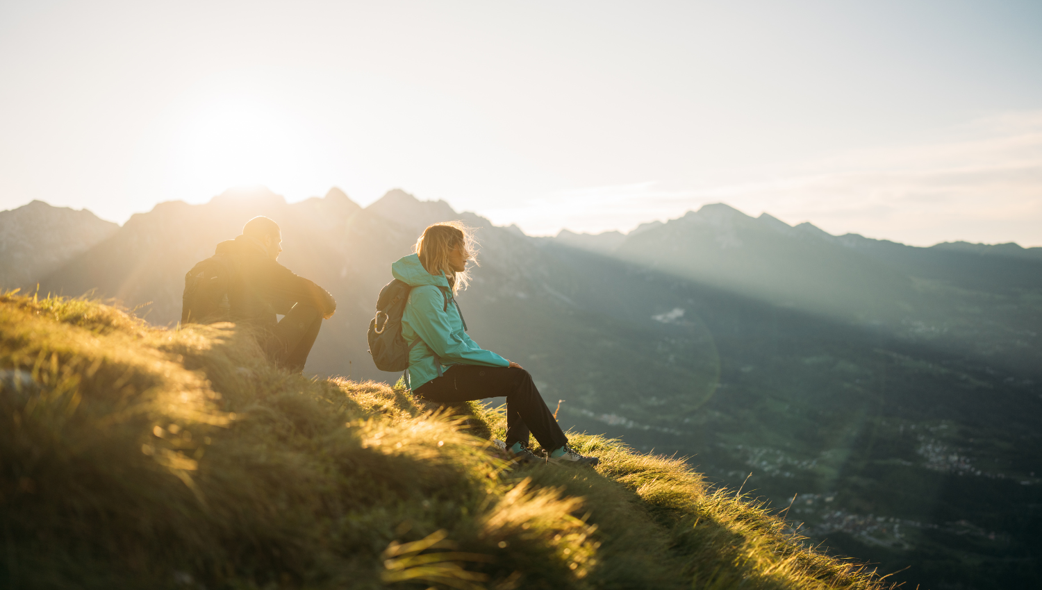 Due persone sedute sull’erba in cima a una montagna al tramonto, con zaini da trekking e vista panoramica sulle valli sottostanti