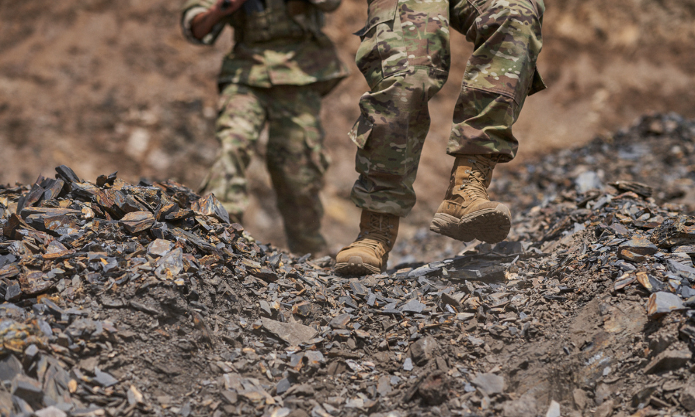 Two soldiers in camouflage uniforms and Garmont tactical boots walking over sharp, dark rock debris on rugged terrain