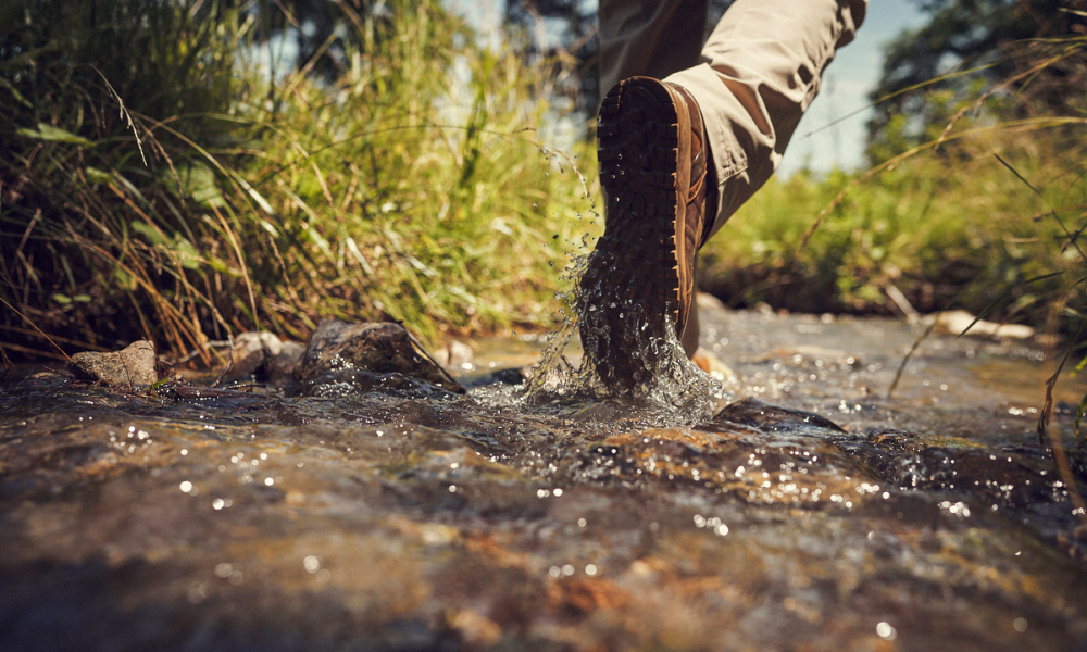 Close-up of a Garmont Tactical boot splashing through a shallow stream, surrounded by grass and rocks