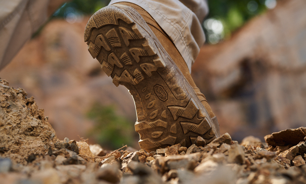 Extreme close-up of the sole of a Garmont tactical boot in motion, kicking up dirt on a rocky trail
