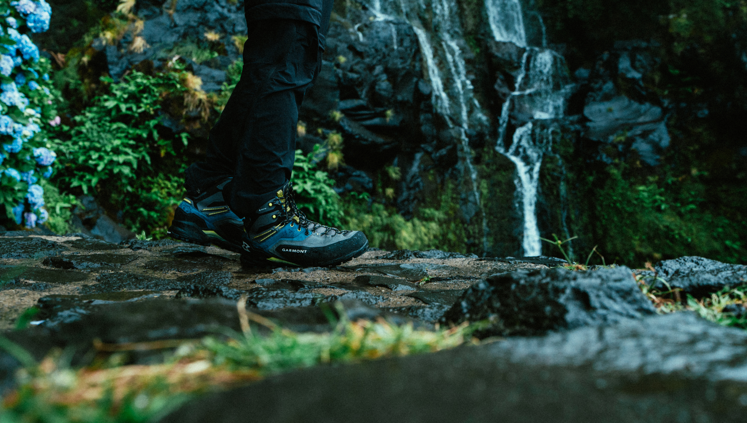 Primo piano di scarponi da trekking Garmont su terreno roccioso bagnato, con una cascata sullo sfondo immersa nella vegetazione.