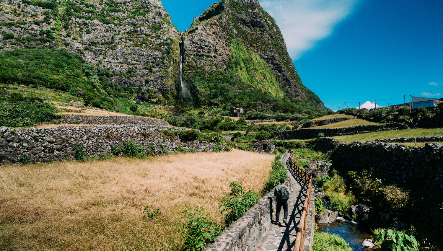 Escursionista che cammina lungo un sentiero in pietra tra muretti a secco, con alte montagne verdi e cascata sullo sfondo.