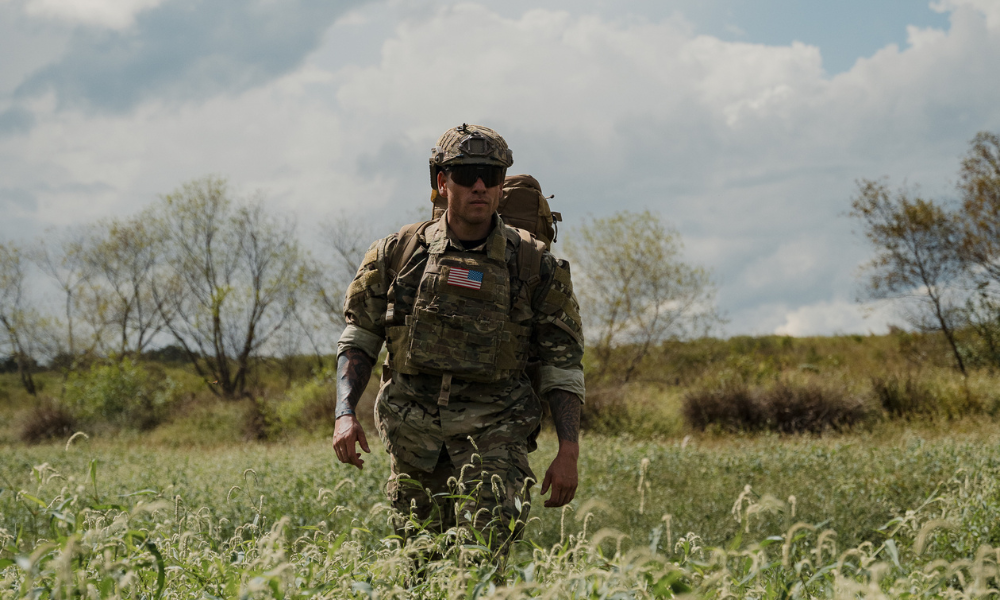 Soldier in full tactical gear walking through tall grass under a cloudy sky, wearing sunglasses and a helmet.