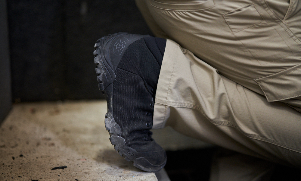 Person kneeling indoors wearing black tactical shoes with reinforced sole and rugged grip.