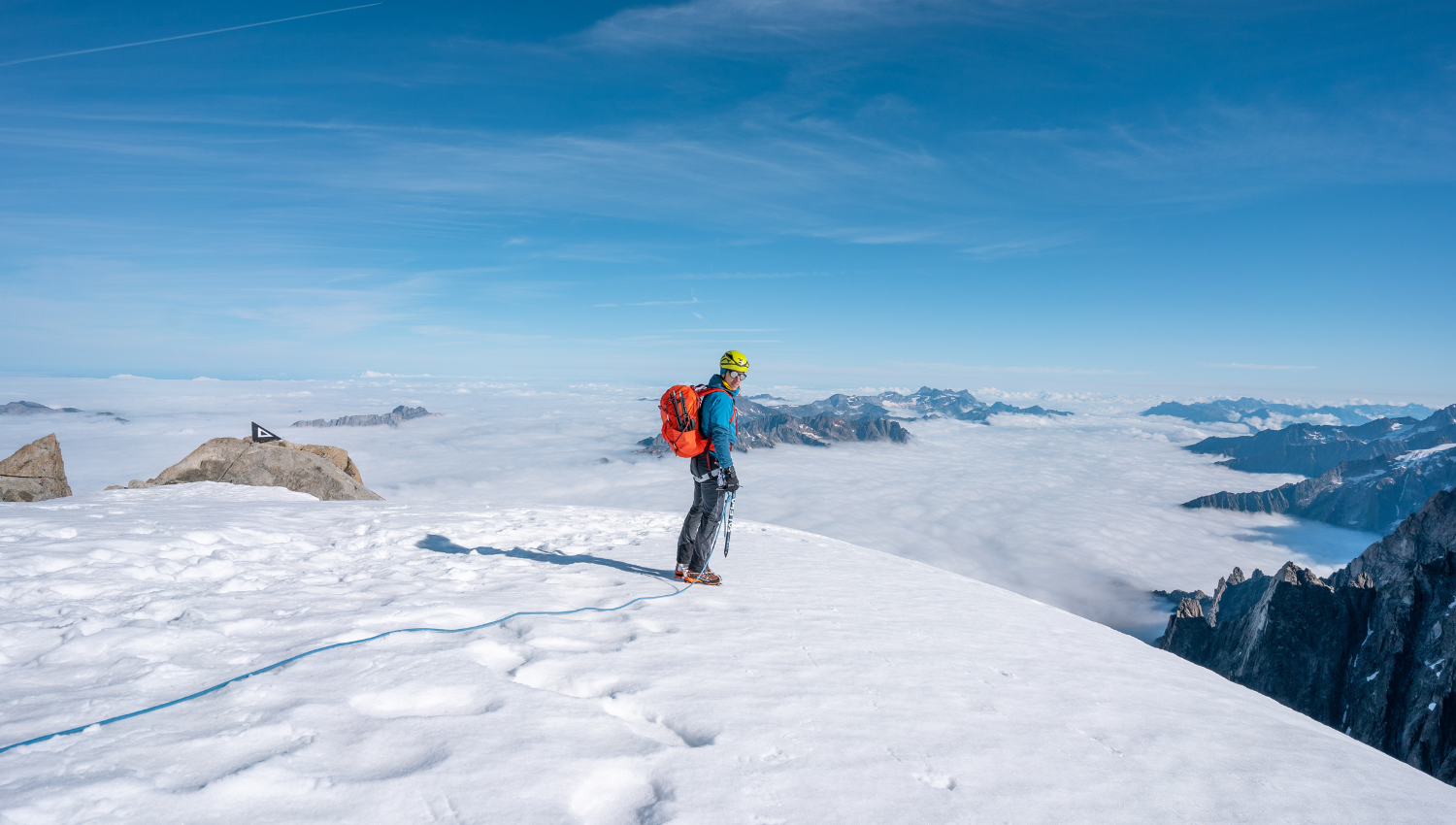Alpinista in cima a una montagna innevata, osserva l’orizzonte sopra un mare di nuvole con le cime delle Alpi in lontananza.