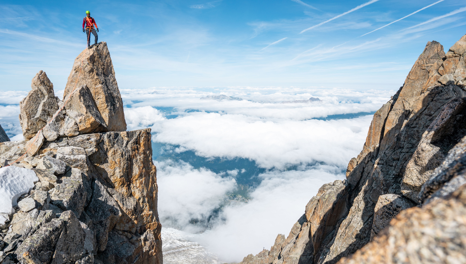 Alpinista in piedi sulla cima di una roccia aguzza, sopra le nuvole, con un panorama montano spettacolare sullo sfondo.
