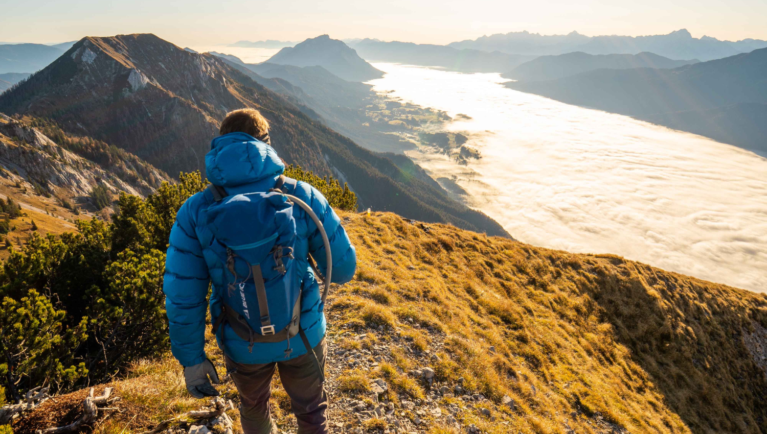 Persona con chaqueta azul observando un valle cubierto de nubes desde una cima montañosa.