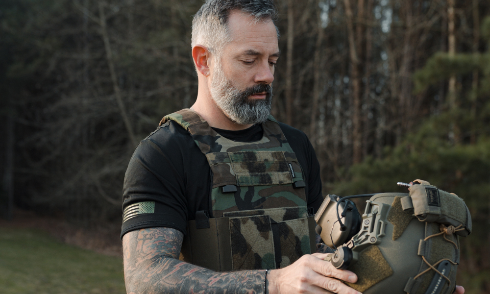 Bearded man in camouflage gear examining a tactical helmet, standing in an outdoor wooded environment.