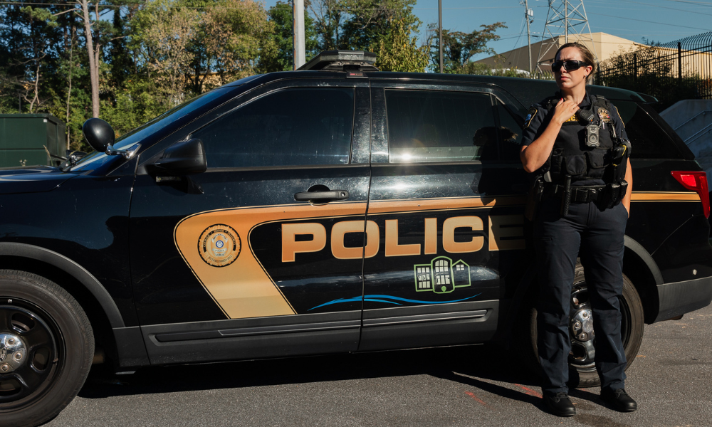 Police officer in uniform standing next to a black and gold patrol SUV, speaking into a shoulder radio microphone.