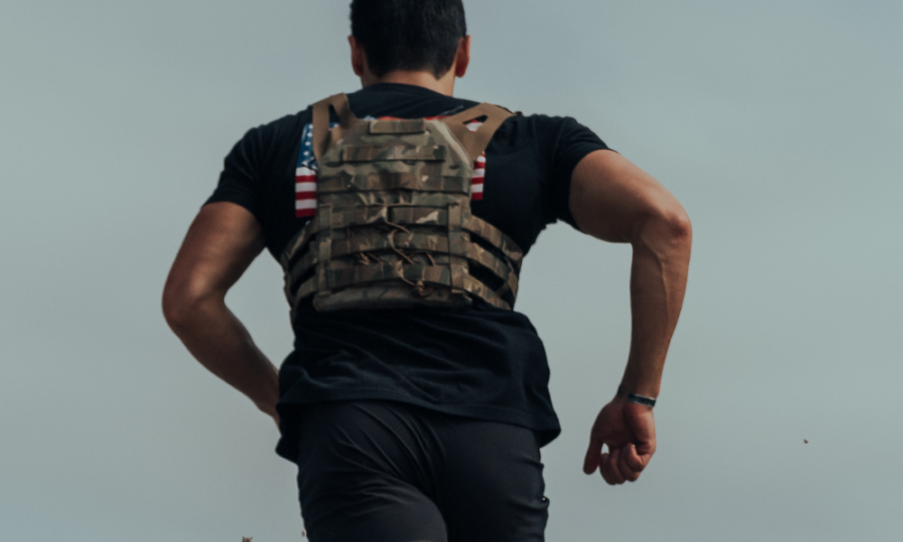 Man running uphill wearing a tactical vest with two American flag patches on the back, captured from behind.