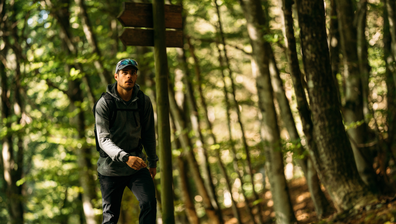 Giovane escursionista con cappellino e zaino in una foresta rigogliosa vicino a un cartello di legno, suggerendo un trekking o un'avventura all'aperto.