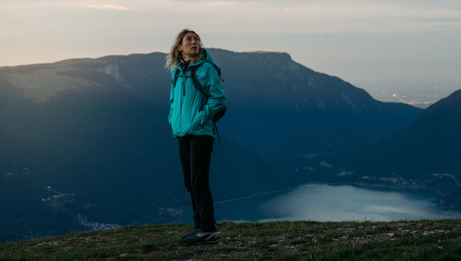 Donna con giacca azzurra e zaino in piedi su un’altura, con lo sguardo rivolto verso l’alto e un lago sullo sfondo tra le montagne al tramonto.