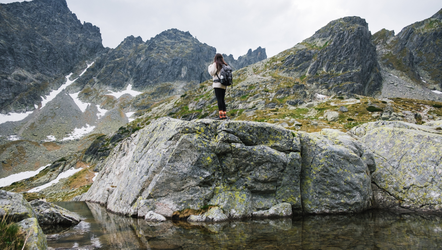 Donna con zaino nero che osserva il panorama da una grande roccia in mezzo alle montagne rocciose, con chiazze di neve e cielo coperto.