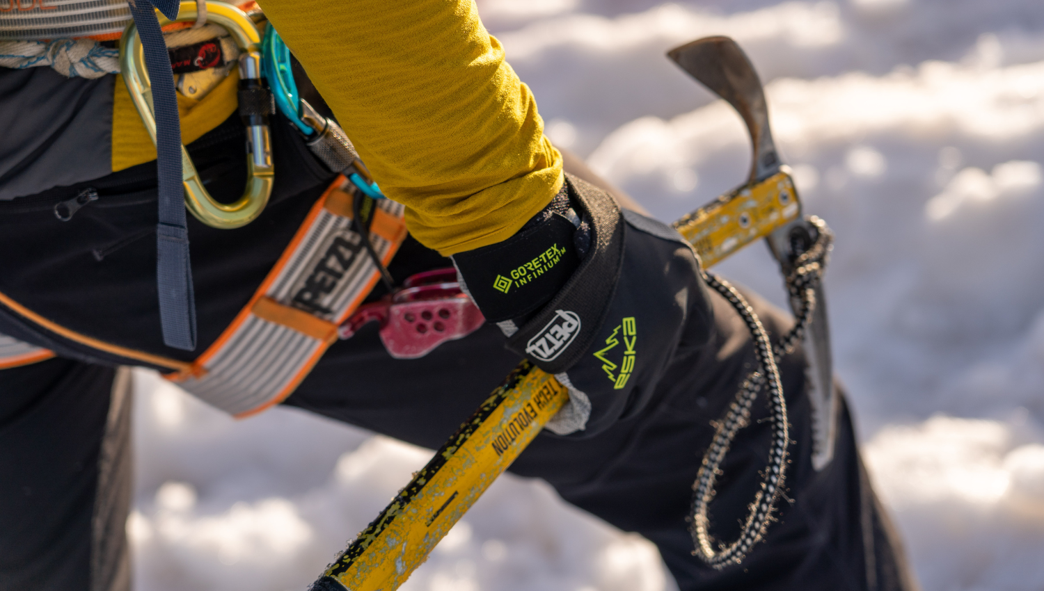 Dettaglio di un alpinista con guanti tecnici, attrezzi da ghiaccio e imbrago, su sfondo innevato.