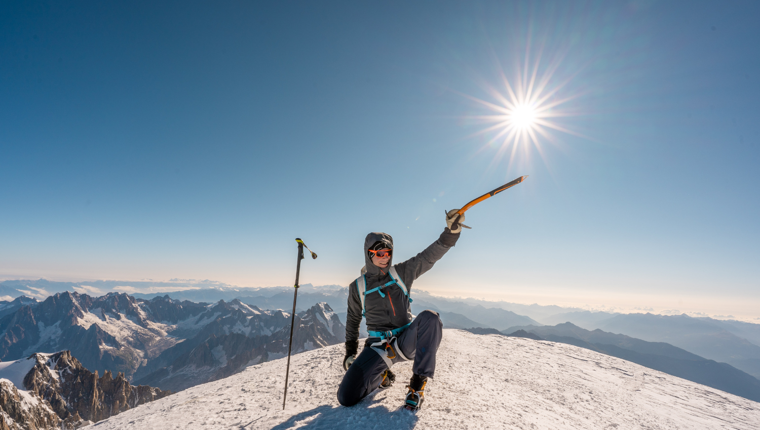 Alpinista in cima a una montagna innevata che solleva la piccozza verso il sole, con vista panoramica sulle Alpi.