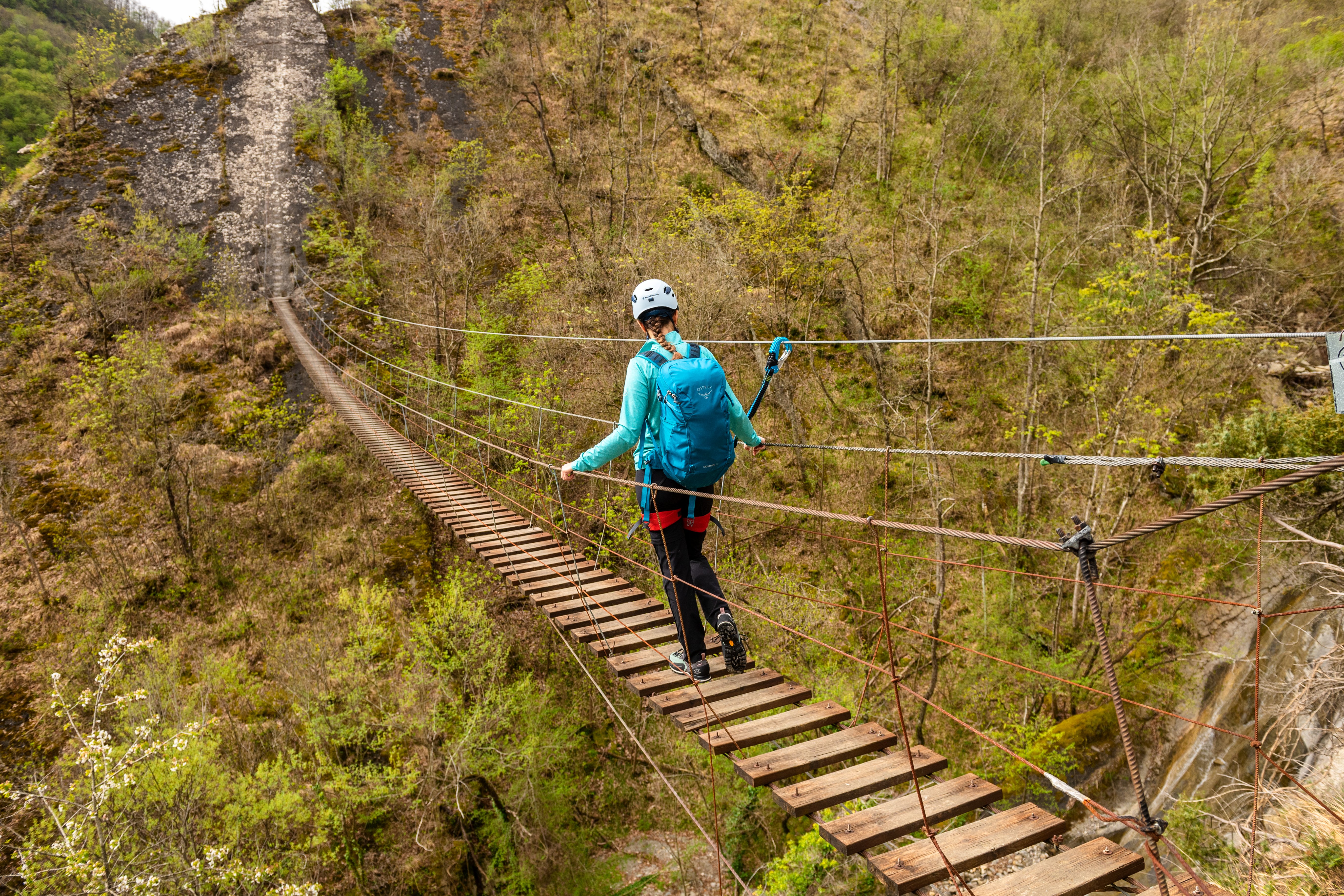 Escursionista con casco, zaino e imbracatura che cammina su un ponte sospeso in legno tra due pareti rocciose, immerso nella vegetazione.