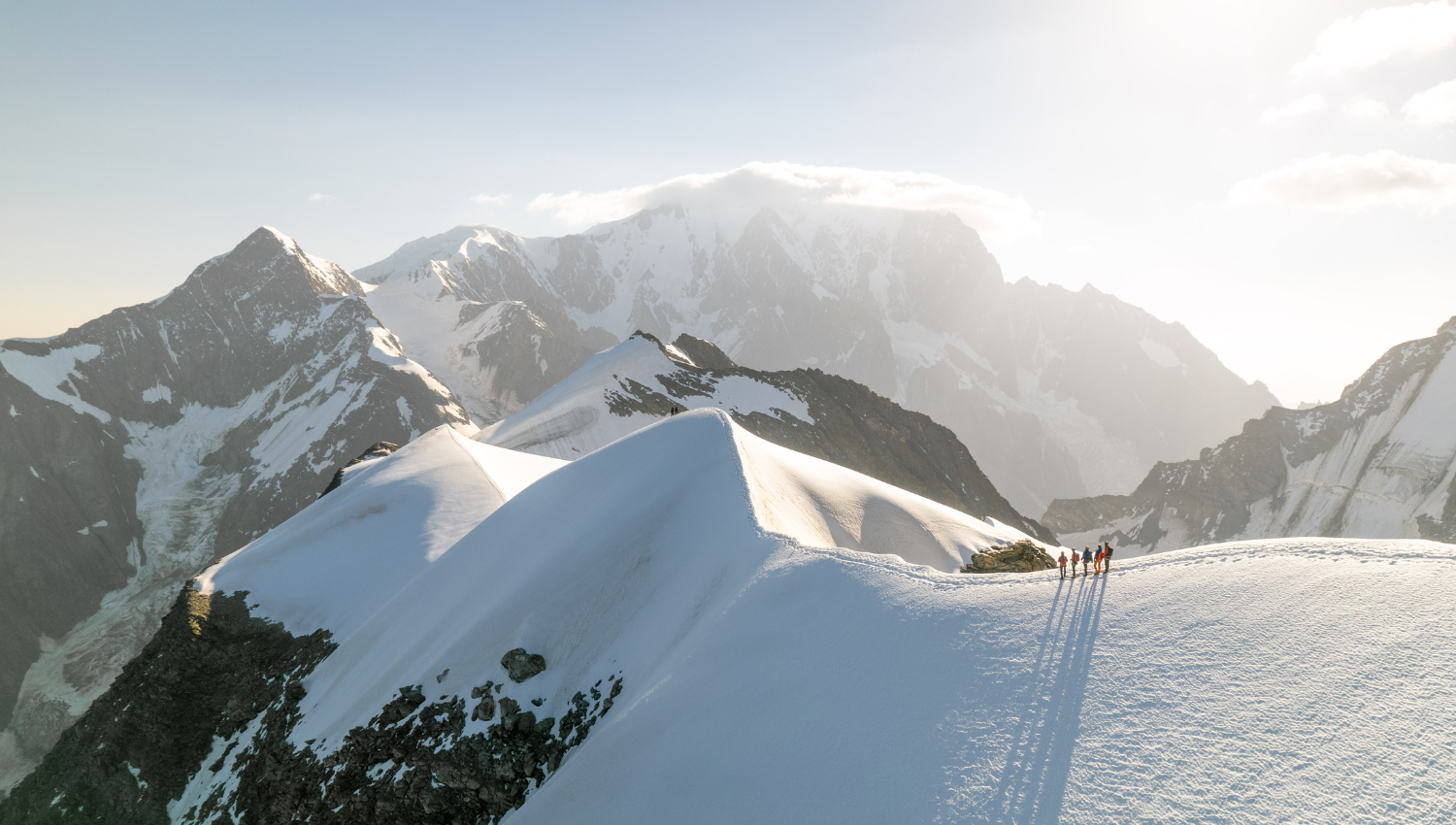 Gruppo di alpinisti che percorrono una cresta innevata, con il Monte Bianco sullo sfondo illuminato dal sole.