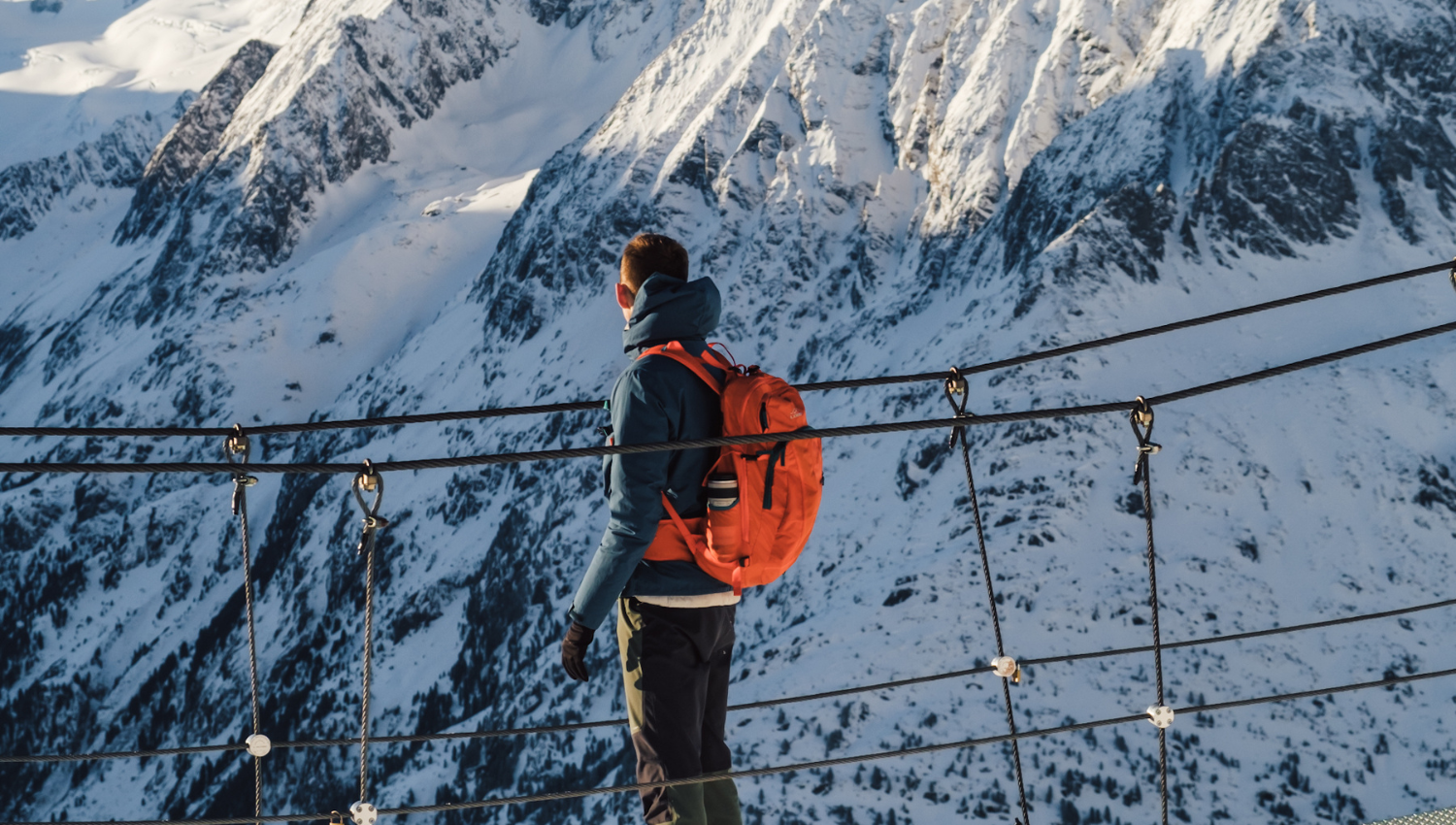 Persona con zaino arancione su un ponte sospeso, osserva una montagna innevata.