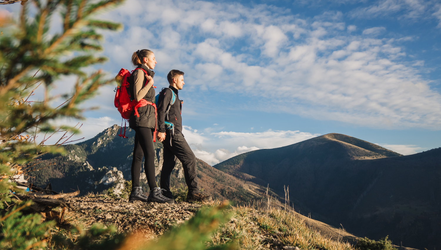 Due escursionisti con zaini rossi osservano il panorama da un crinale montano, sotto un cielo sereno punteggiato da nuvole.