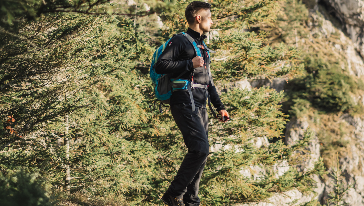 Persona in abbigliamento da trekking cammina tra gli alberi con uno zaino blu, circondata da pini verdi.