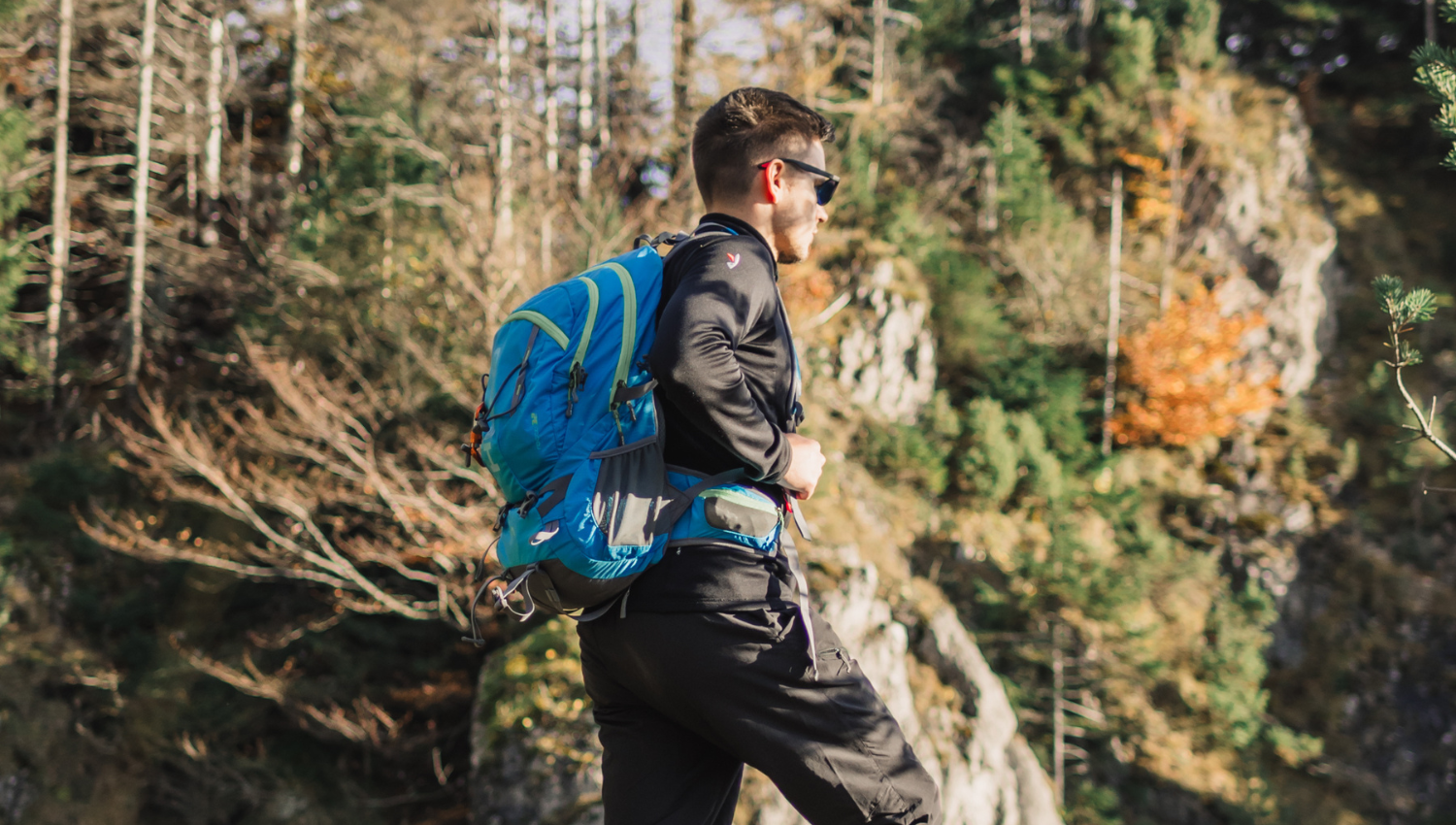 Excursionista con mochila azul caminando entre árboles en un sendero de montaña.