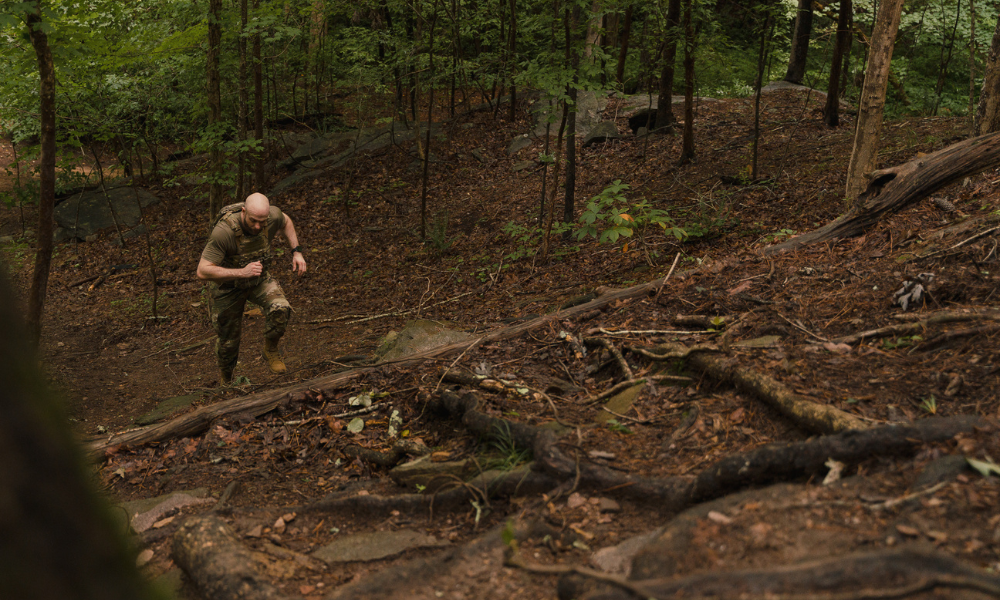 Man in tactical gear running uphill through dense forest terrain.