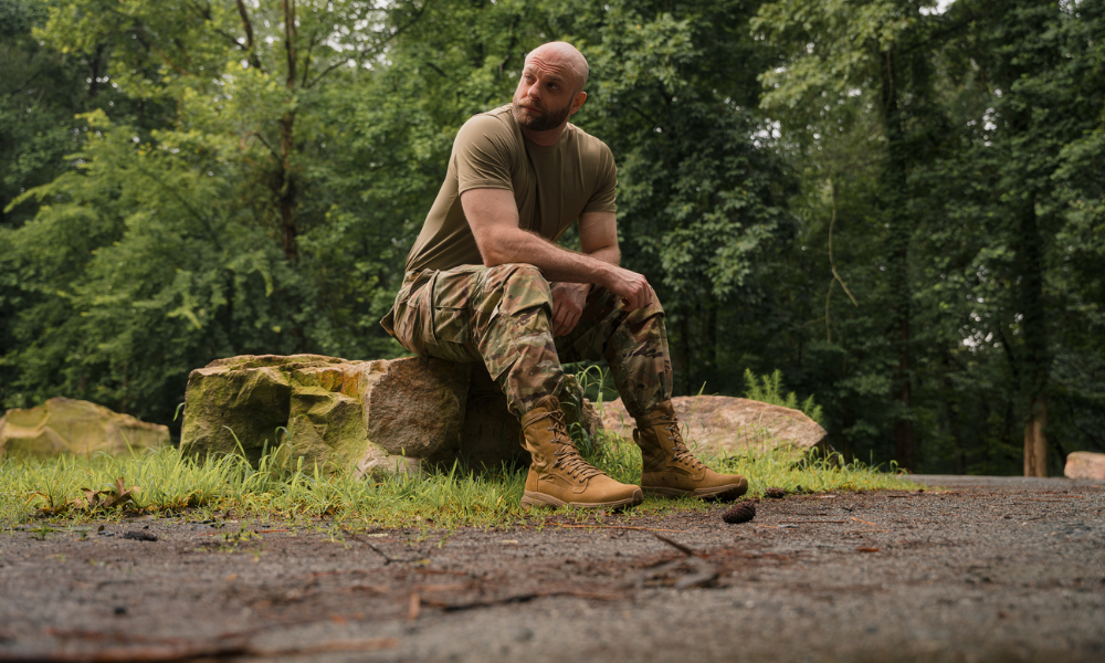 Man in camouflage pants and tactical boots sitting on a rock in a forest setting.