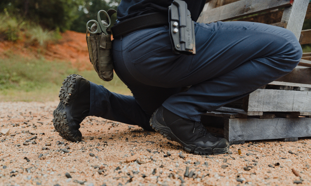 Close-up of tactical boots and holstered pistol as a person kneels on gravel.