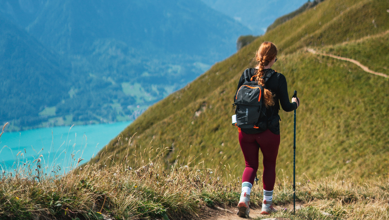 Escursionista di spalle che cammina lungo un sentiero erboso con vista su un lago d'Iseo dalle acque turchesi e montagne circostanti.