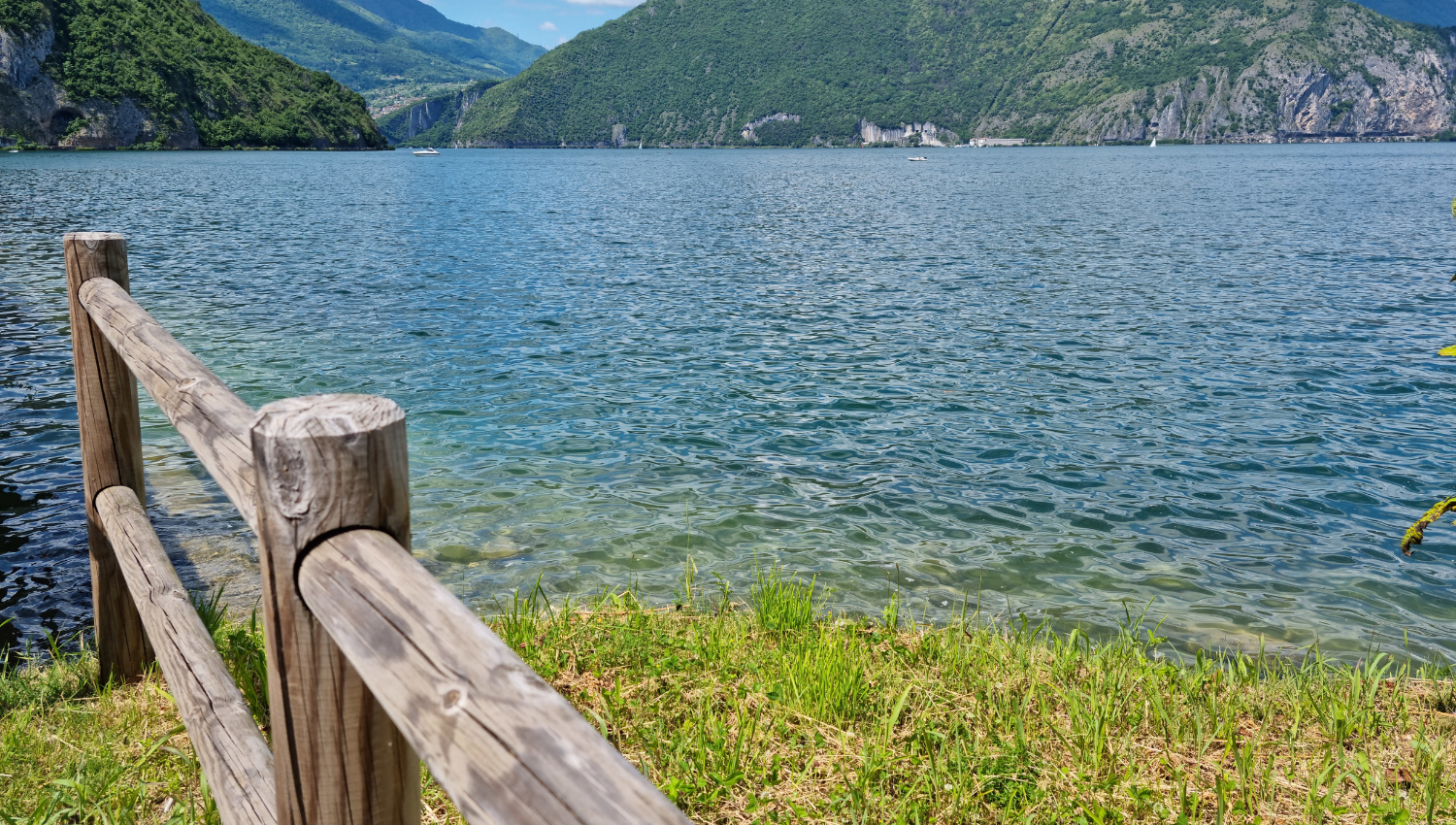 Vista panoramica sul lago d’Iseo con acque cristalline, montagne verdi sullo sfondo e una staccionata in legno in primo piano.
