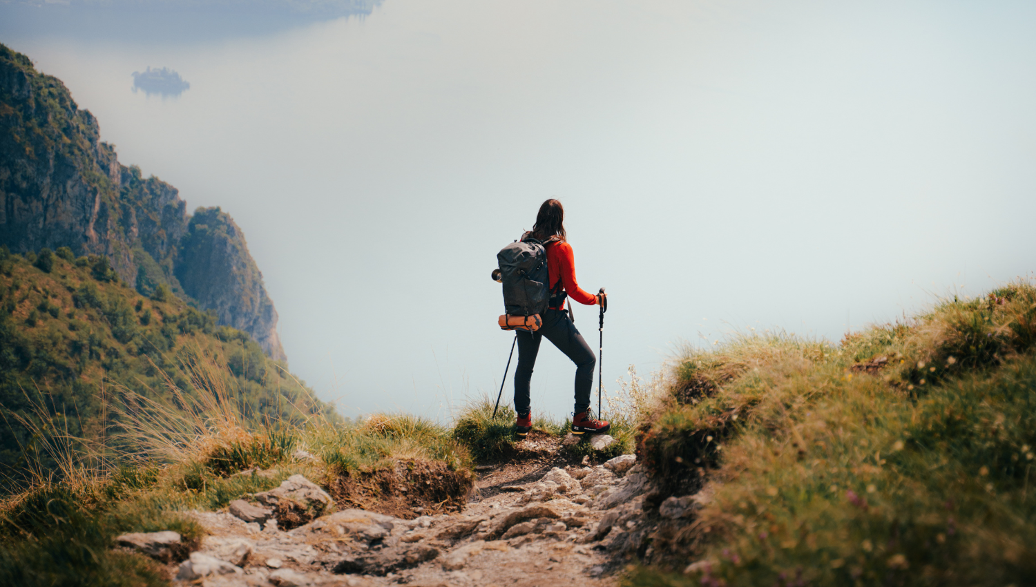 Escursionista in cima a un sentiero roccioso con vista mozzafiato sul lago, equipaggiata con zaino e bastoncini da trekking.