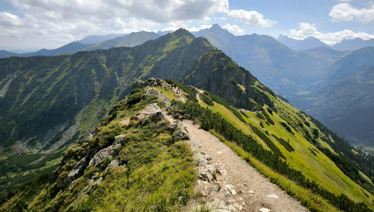  Sendero de cresta en lo alto de una cadena montañosa, con vistas panorámicas de cumbres verdes y cielos parcialmente nublados.