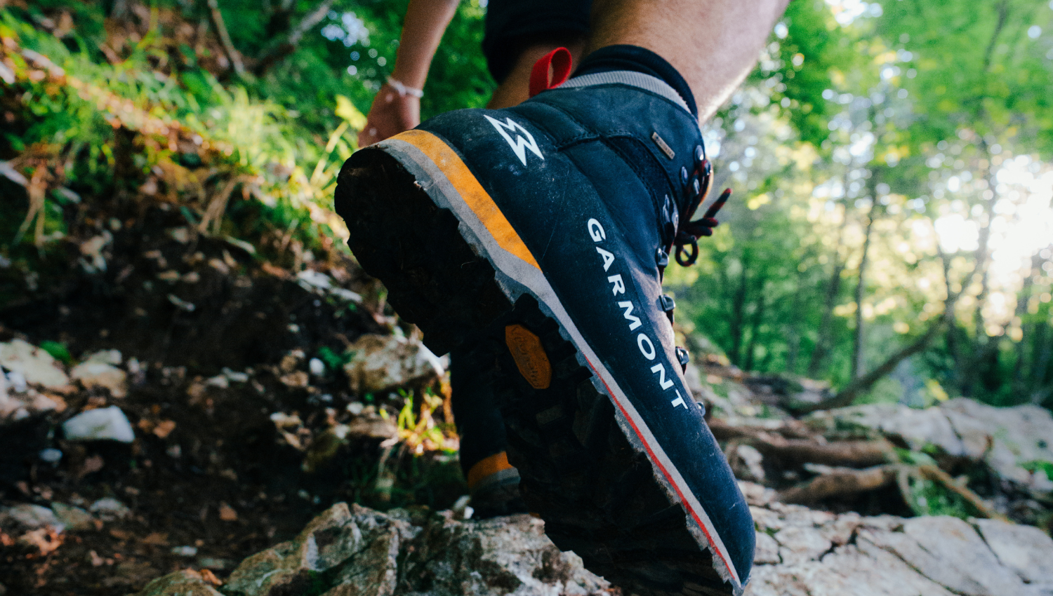 Primer plano de una bota de montaña Garmont en acción sobre un sendero rocoso en medio del bosque.
