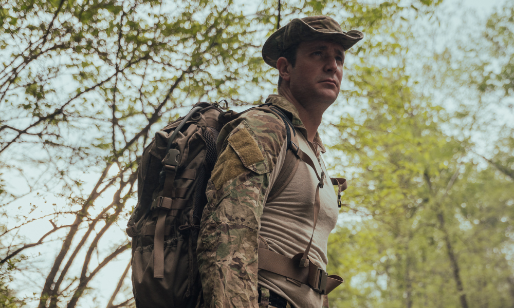 Male soldier wearing camouflage uniform and wide-brim hat, standing alert in a forest with a large tactical backpack and chest harness.